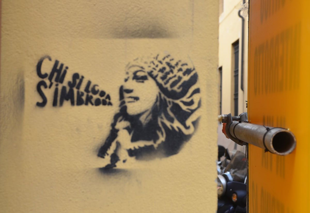 black stencil of a woman's head in profile, long hair, wearing a hat, words coming out of her mouth, shape of words is a megaphone. Words say " Chi si loda s'imbroda"