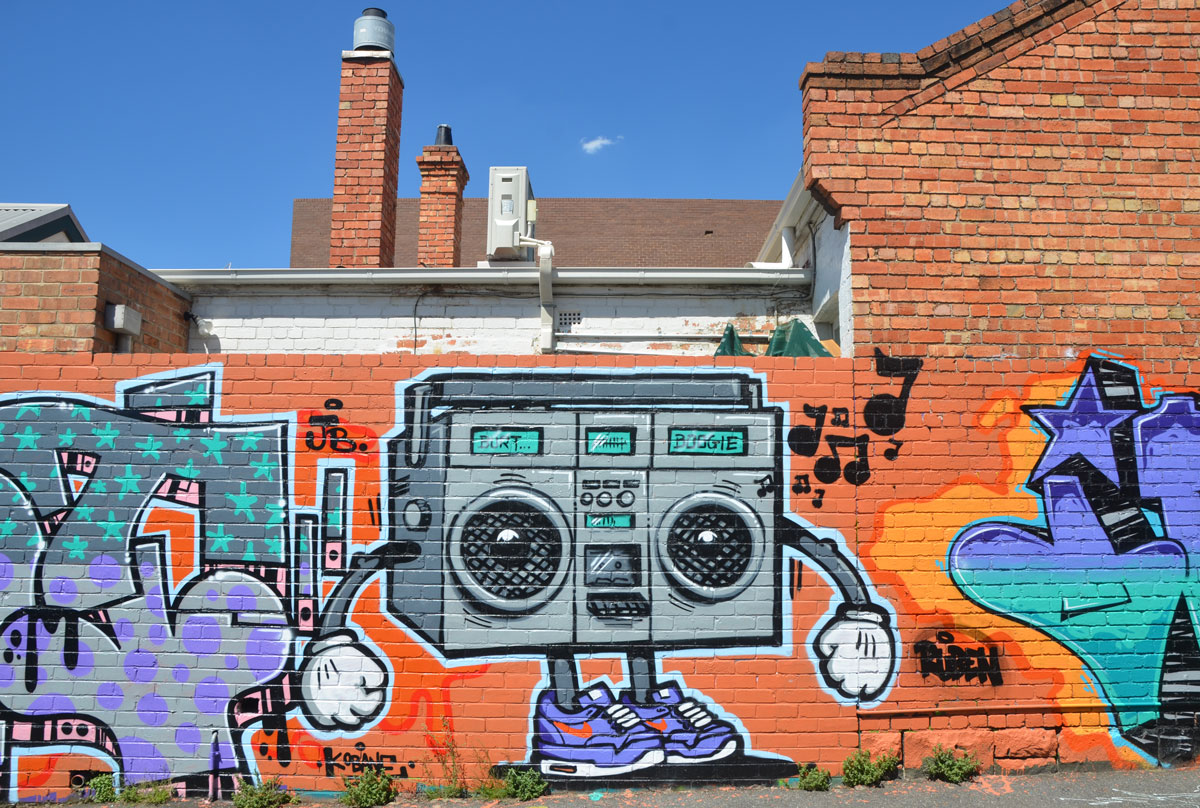 a street art painting of a ghetto blaster with speakers that look like round eye, tiny legs with sneakers and arms with big white hands.