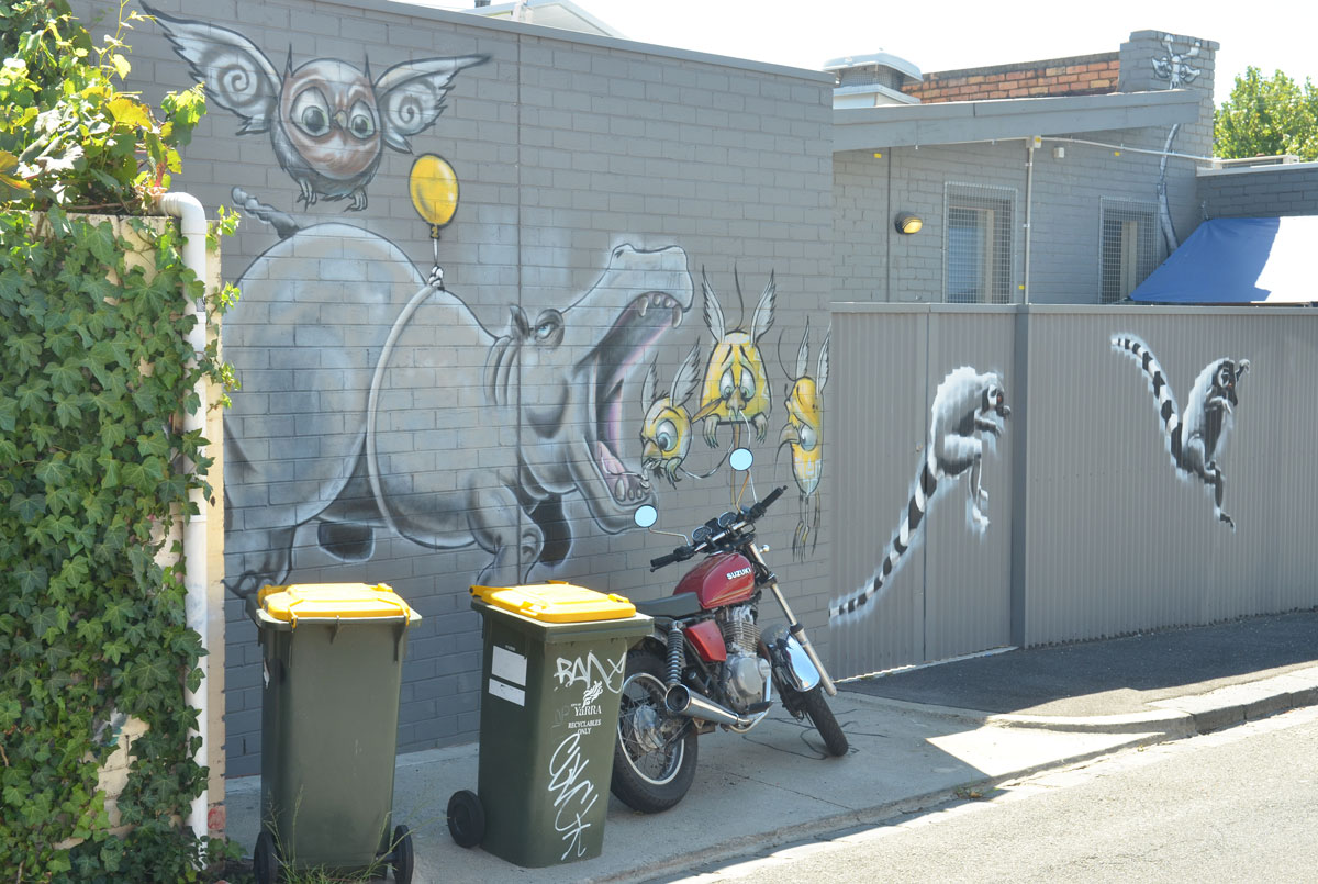two garbage bins and a motorcycle in front of a mural on a grey wall. Mural is of a large rhino with its mouth open, trying to catch birds in its mouth, two leaping lemurs and leaping away from the rhinocerous.