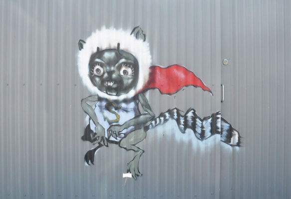 street art painting of an animal with a black and white striped tail, wearing a fuzzy white band around its head, and a red cape. Also wearing a shirt with a big S on it.
