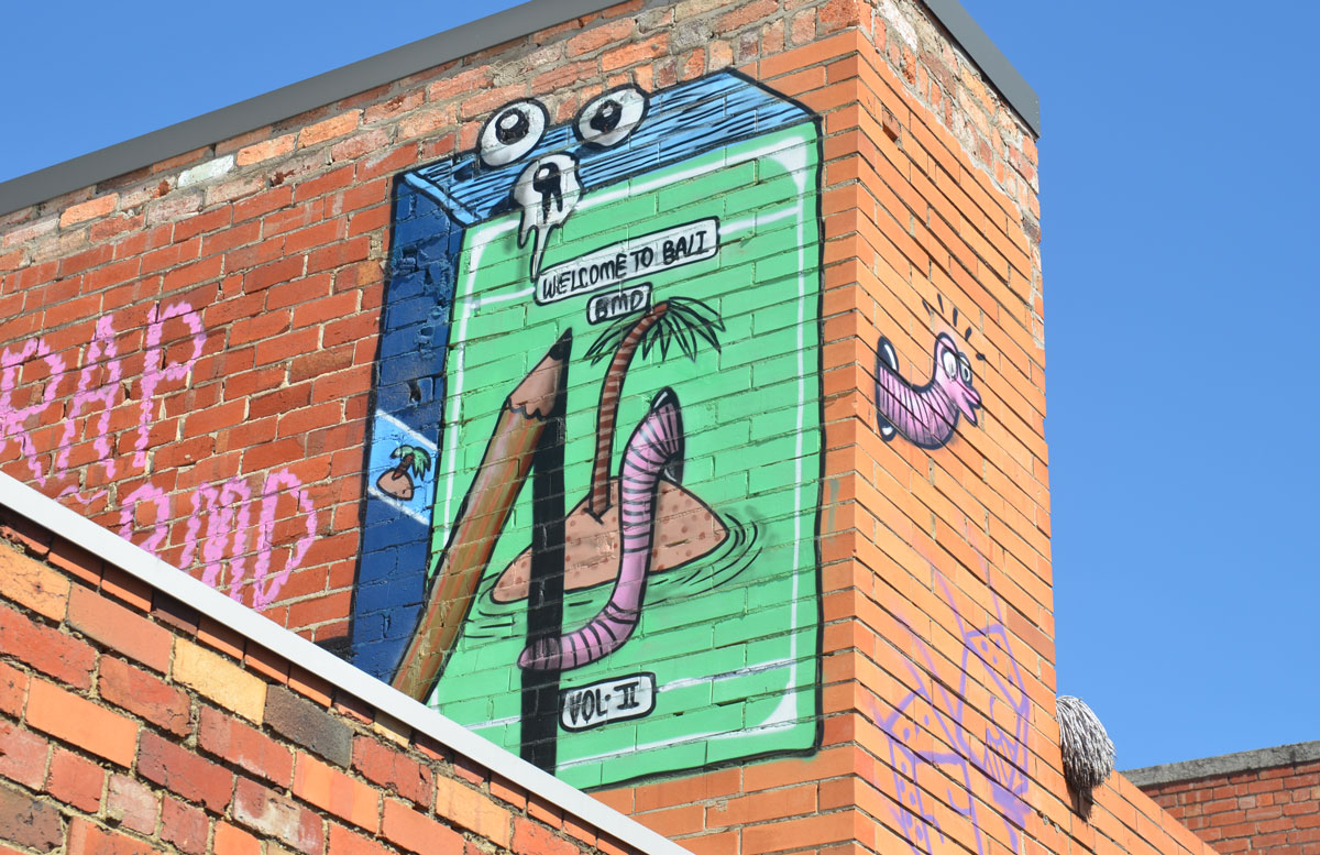 muralby BMD on the upper level of a red brick building. Cigarette box shaped box, front is green with a picture of an little island with one little palm tree and a large earthworm coming out of the sand. a large pencil is seen drawing the picture