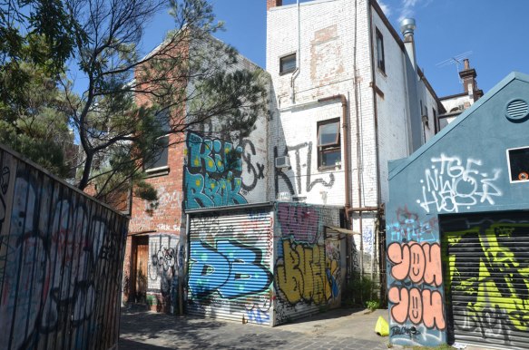 the backs of buildings in a lane. An old wood fence, with a tree behind it. A taller white building among some shorter buildings. sunny day, blue sky, Melbourne Australia.