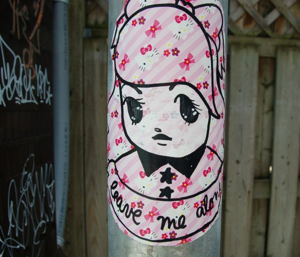 Starchild Stela character, a girl with shoulder length and slightly curly hair, sticker on a grey metal pole, pale pink background with little pink bows pattern, with the words 'leave me alone' written under it