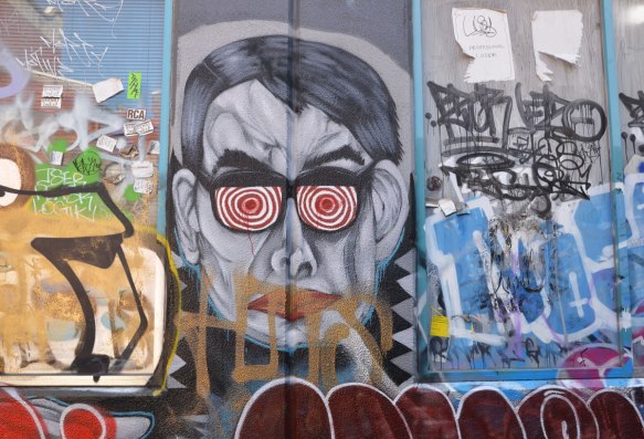 graffiti and street art on a wall, some scrawls and scribbles but also one man's face, wearing glasses with red and white concentric circles instead of lenses
