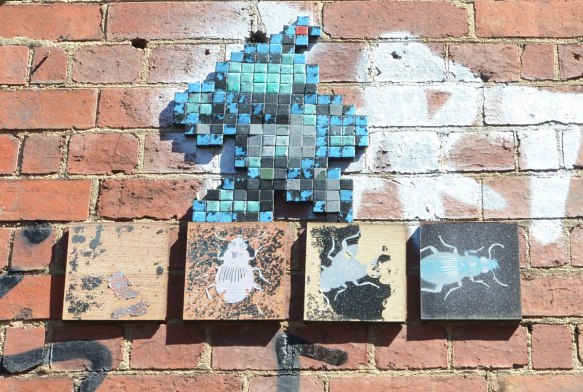 four square pieces of wood mounted on a wall, each with a picture of an insect. Above it is a creature, walking on two legs, made of black and blue mosaic tiles
