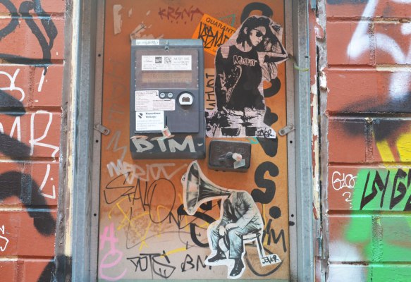 2 paste ups on a boarded up space on a wall. One is a person with a black t shirt with the word Misfits on it. The other is a man sitting on a stool but instead of a head he has the speaker part of an old gramophone