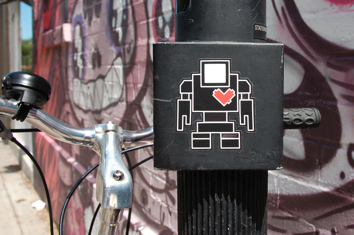 a black lovebot sticker on a black pole with a bike parked against that pole and a pink and purple mural of faces behind in the background