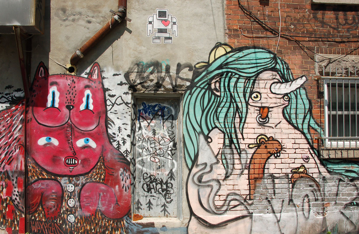 a small lovebot wheatpaste on a wall above some street art painted creatures, one cat like animal in red and one pinkish creature with long green hair.