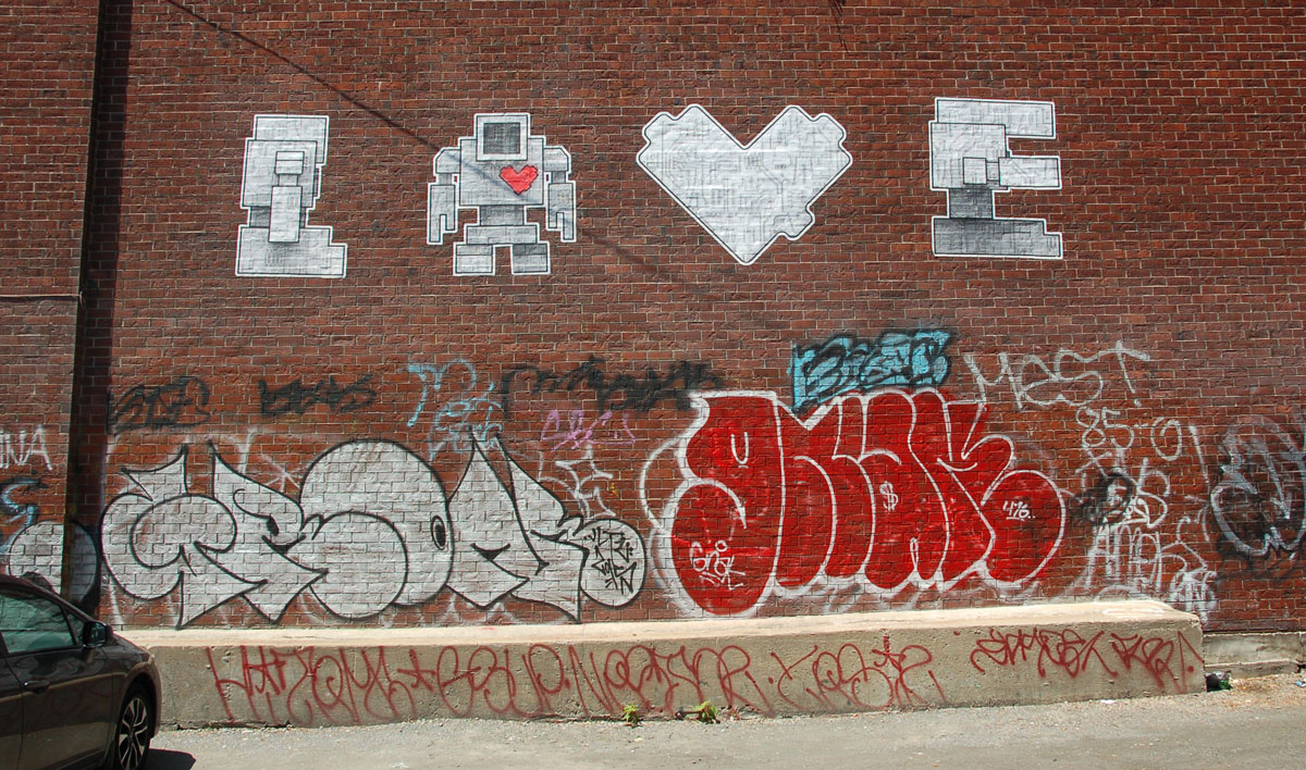 four large wheatpaste pieces high on a brick wall. Lovebot in profile facing right makes an L, then a lovebot facing forward for O, then a lovebot heart for V and finally another lovebot in profile, with arm sticking out, to make E.