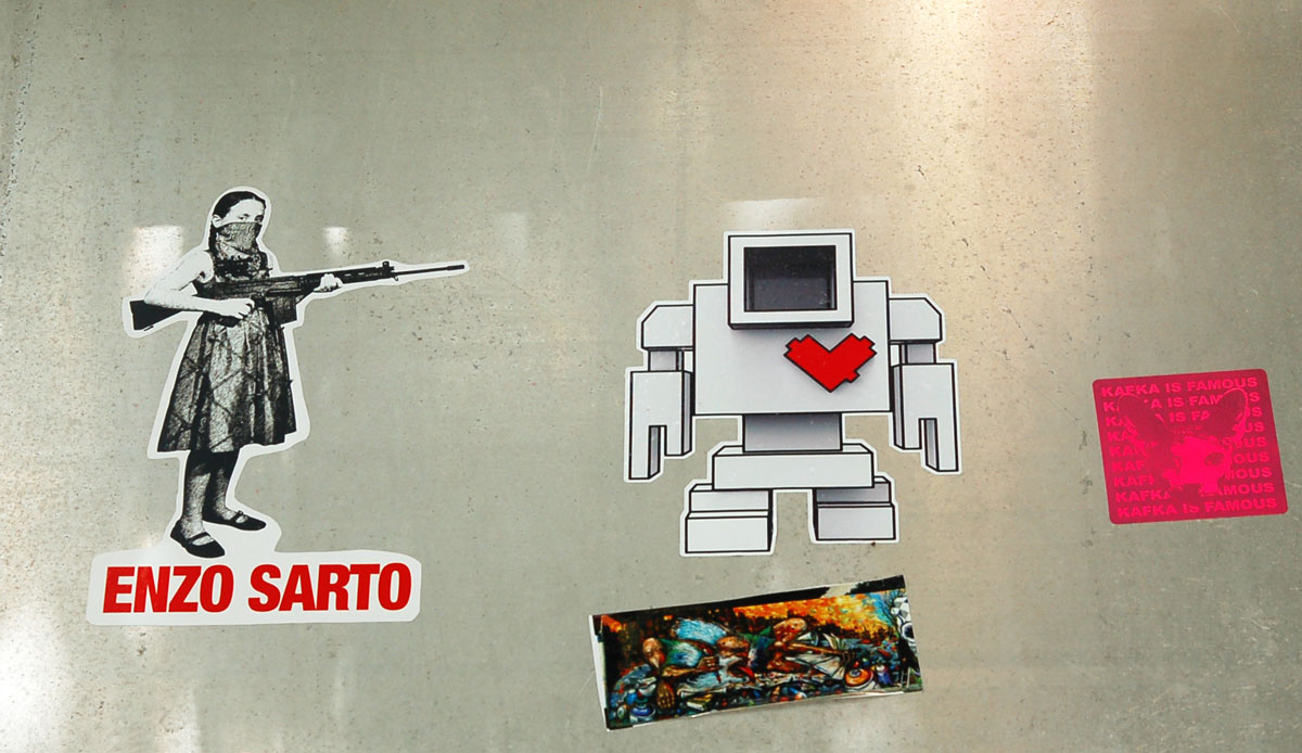 A light grey lovebot sticker beside a sticker of a girl with bandana over the lower part of his face and she is pointing her rifle at lovebot. The sticker of the girl is by Enzo Sarto and his name on it.