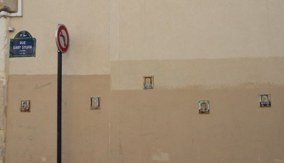 On a light brown stucco wall, sign for rue Gaby Sylvia, as well as 5 small portraits frames in tiny mosiac tiles 