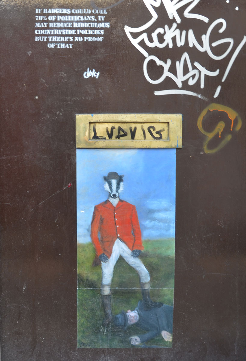 street art painting of a hunter in red jacket but with badger face standing over a person who is lying on the ground, one leg of badger person is on the other person's back