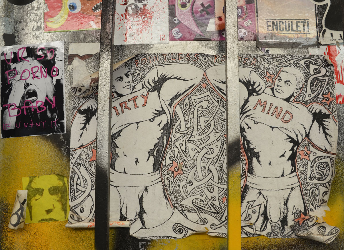 a group of pasteups on a wall. the largest one is of two guys in their underwear pulling their shirts up to reveal their stomachs and chest. One man has 'dirty' written on the shirt and the other has 'mind' written on his.