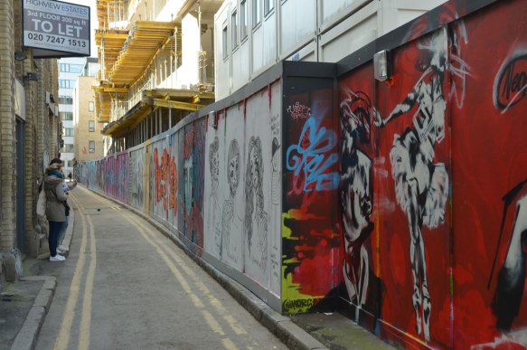 view of a narrow London Street with street art covered construction hoardings down one side, two young women taking photos of the hoardings on the left. New building under construction in the background. 