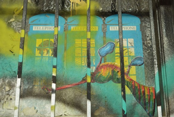 street art, three blue British style phone booths behind bars, with a creature in front of them 