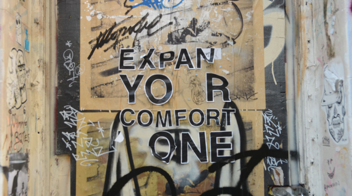 words on a graffiti covered wall that once said Expand your comfort zone, but some of the letters are missing and others are starting to peel. 