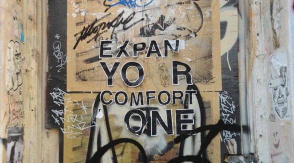 words on a graffiti covered wall that once said Expand your comfort zone, but some of the letters are missing and others are starting to peel. 