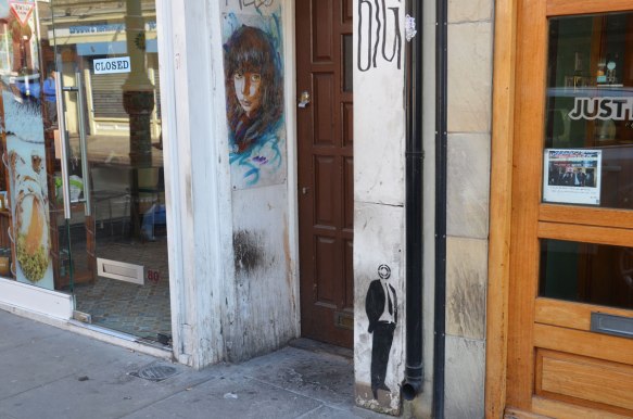 a very small black stencil of a man with a target as a head, standing on a wall, with feet at sidewalk level . Near him is a woman's face painted on the wall of a doorway 