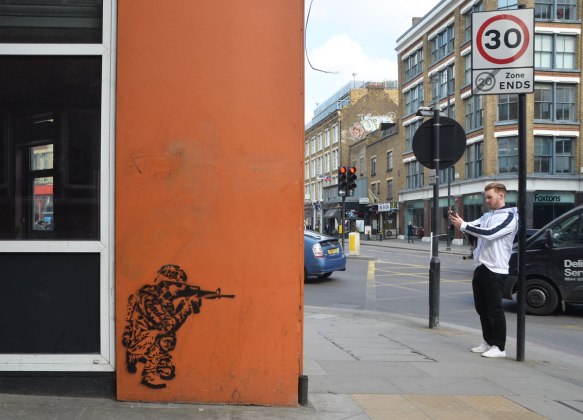 street art piece of a person squatting beside a building, shooting an automatic weapon towards the edge of the building. A young man is taking a picture with his phone on the other side of the building. 