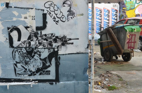 street art piece of a person squatting beside a building, shooting an automatic weapon towards the edge of the building. A trash can and car are beside the building 