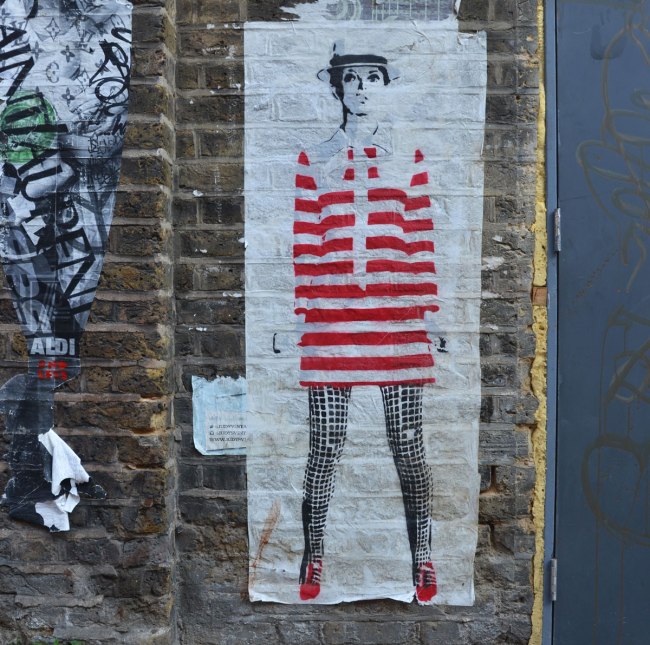 large paste up of woman standing, looks like Liza Minelli in red and white striped long top and black leggings, red shoes, on a brick wall 