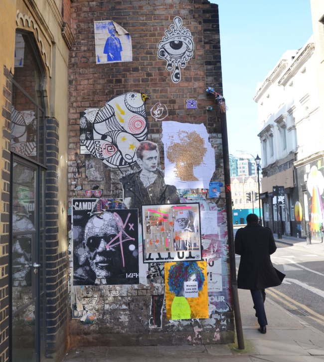 a man walks past a wall that is covered with stickers and paste ups graffiti and street art in Shoreditch London