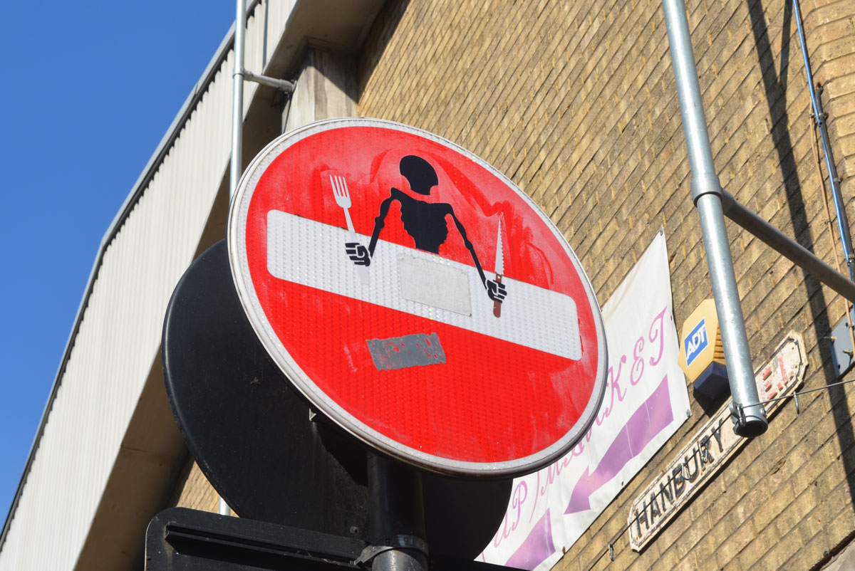 altered red and white no entry street sign - the white bar is a table, black figure sitting at the table with hands on the table. cutlery in hands, no food on the table 