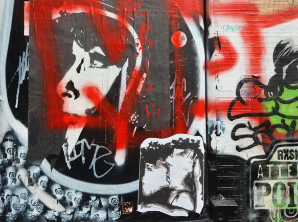 graffiti stencils and paste ups on a wall, a woman's head in profile, a couple kissing, red spray scrawls 
