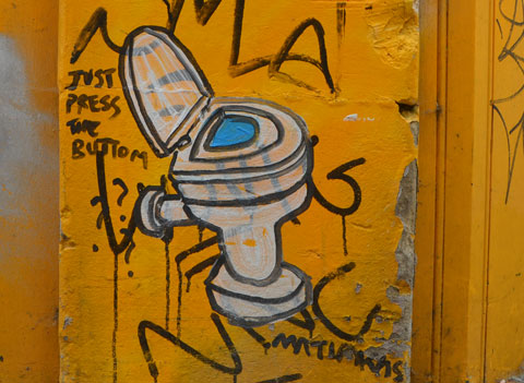 graffiti drawn on a yellow wall, a toilet with the lid up and words that say Just press the button 