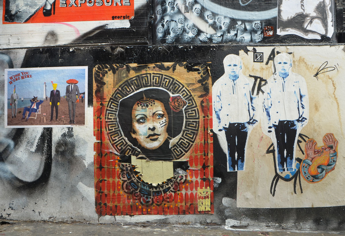 graffiti and street art on a wall, including a beach scene by face the strange, a woman, and two twin men. 