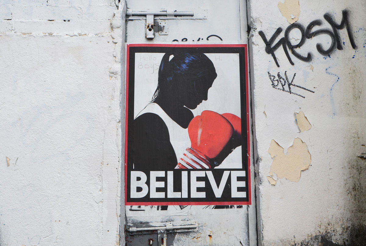 poster on a grubby dirty white wall of a woman in profile, black with white top and red boxing gloves held in front of her chest. The word believe is written in capital letters under her