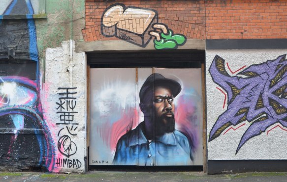 a street art painting of a slice of bread with green feet is above a street art painting by deph of a black man with a beard and a brown cap