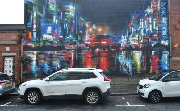 large mural of streets and car traffic at night with lights shining on wet streets, two white cars parked in front. 
