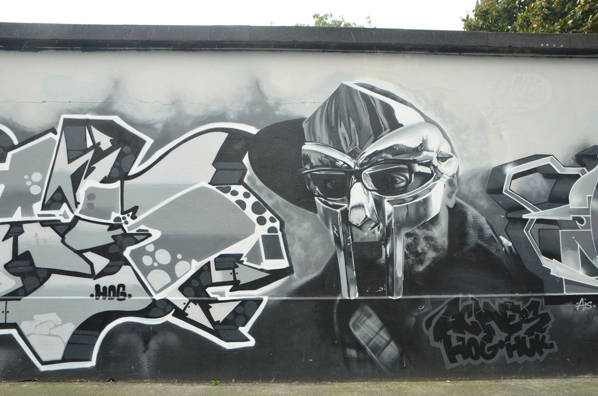 grey tones mural of a man in a mask with text street art on either side of his head and shoulders, words say aches hog huz 