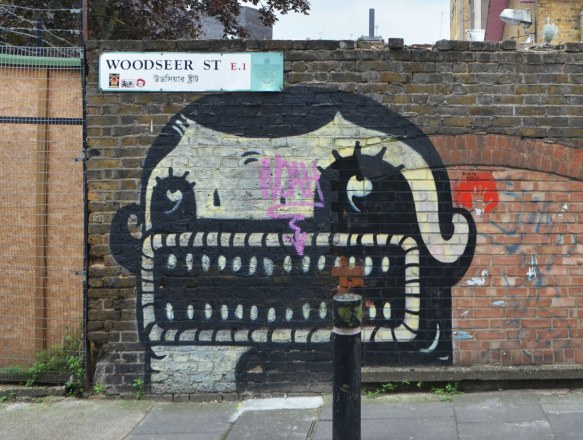 painting by street artist plin of a black and white face that is mostly mouth - a large rectangular mouth with a lot of little teeth, two eyes and only a small bit of black hair. On a brick wall by a sign (street sign) that says Woodseer St. E.1. It also has the name in arabic. 