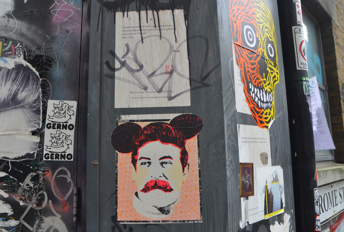 many pieces of street art, including a man wearing mickey mouse ears, a large grinning skull in yellow and orange, and poems written on posters in Italian. 