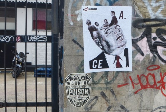 a brick wall beside a metal gate, behind the gate is a motorcycle, on the brick wall are two street art paste ups, a warning poison label and a poster by M. Torannen of a man with the top of head opened up and four people standing inside. 