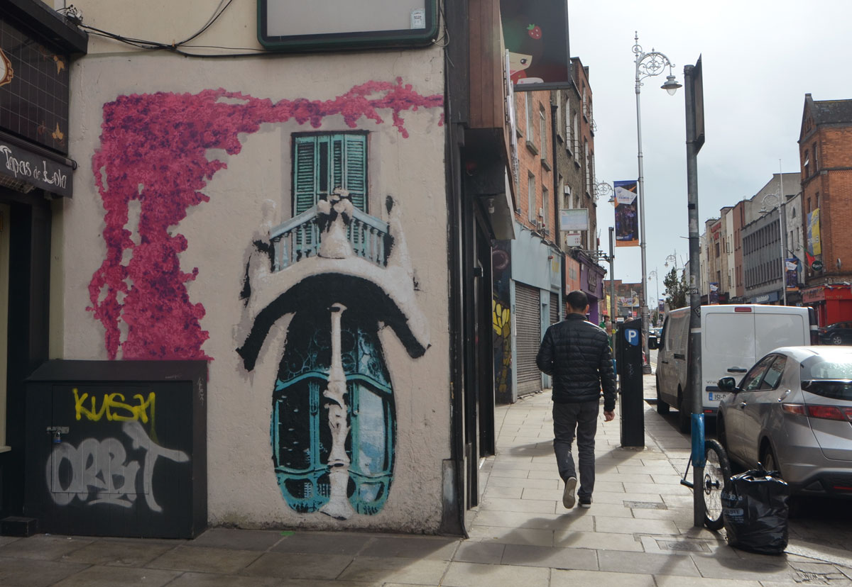 man walking away from the camera, walking past a mural on the side of a shop, pink vine growing upwards, a small round balcony with a window.