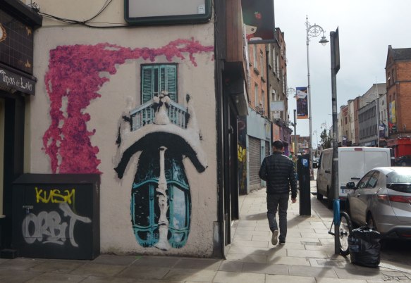 man walking away from the camera, walking past a mural on the side of a shop, pink vine growing upwards, a small round balcony with a window. 
