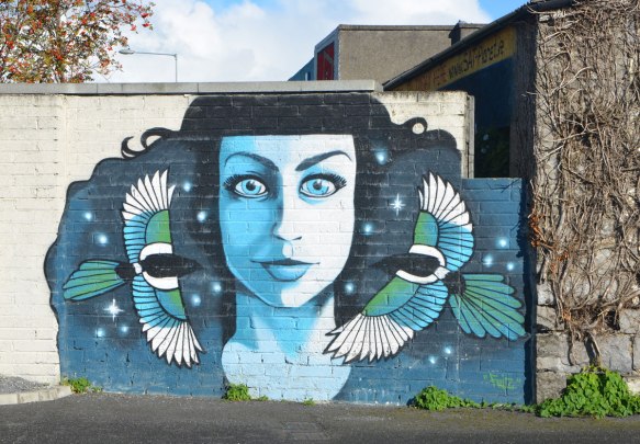 a mural of a woman in blue tones, frontal image, staring straight ahead. Two birds are in flight, one on either side of her, flying towards the center of the picture