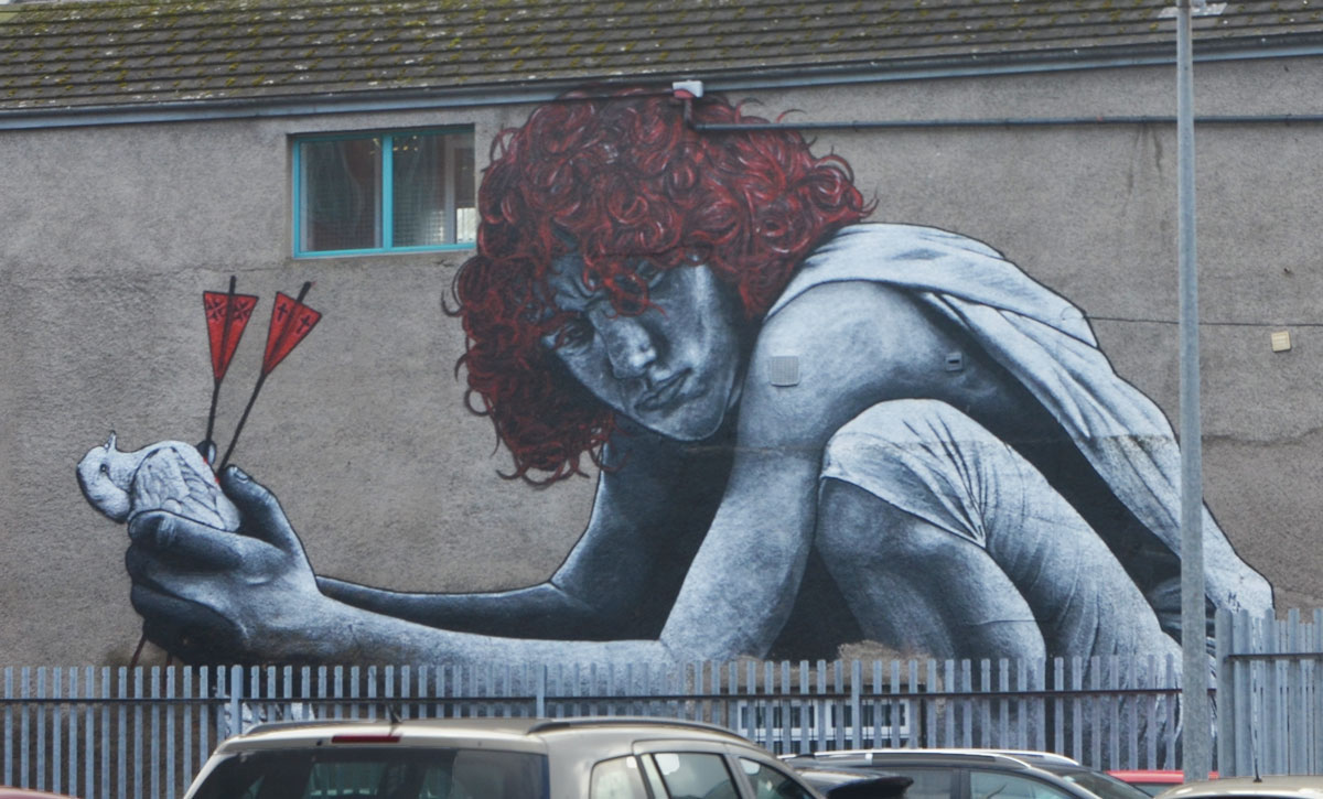 mural of a red headed young man squatting and holding a dead bird in his hands. The bird has two arrows through it, with red ends on the arrows. The mural looks to be on top of a fence and there are cars parked in front of the fence