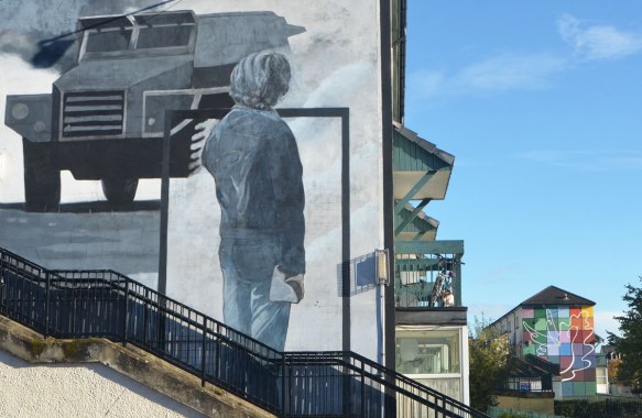 Free Derry mural in Bogside, Derry Northern Ireland, in shades of grey, commemorating Bloody Sunday in 1972 - man with back to viewer watches a tank. Second mural in the background of a white outline of a peace dove over a chequer board design in many different colours. 