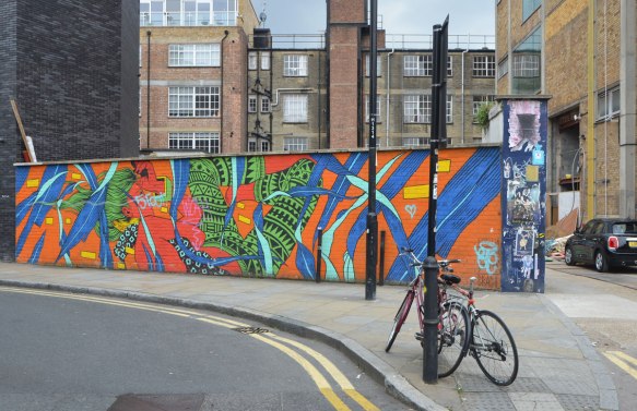 mural by bicicletas on a low brick fence on a street, low rise apartments in the background, a bike parked in front.  Bright and bold colours, red, blue, green and orange mostly, abstracted nature scene