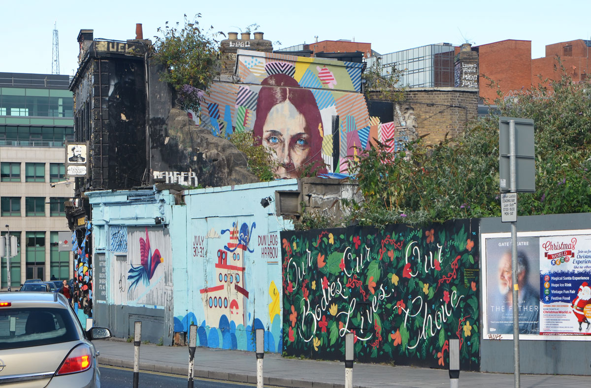 murals on a wall and on the upper storey of a pub behind the fence, a tug boat, fowers, a woman's head 