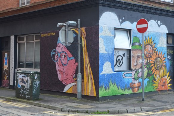 two murals on a corner of a building. One one sideis a head and shoulders of a nun in a yellow habit with black head wear. On the other corner is a leprechaun and some sunflowers. 