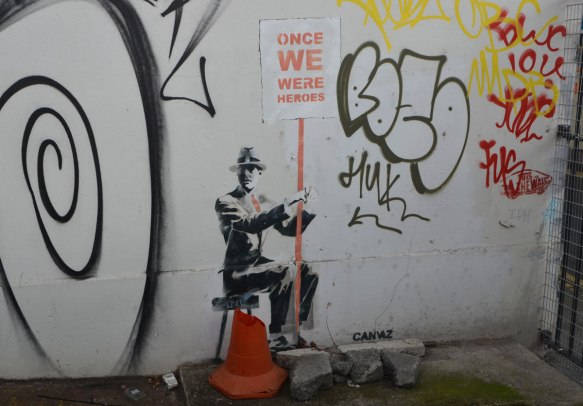 stencil of a man sitting on a stool, wearing a suit and hat, and holding a sign with red letters on white that say Once we were heroes. 