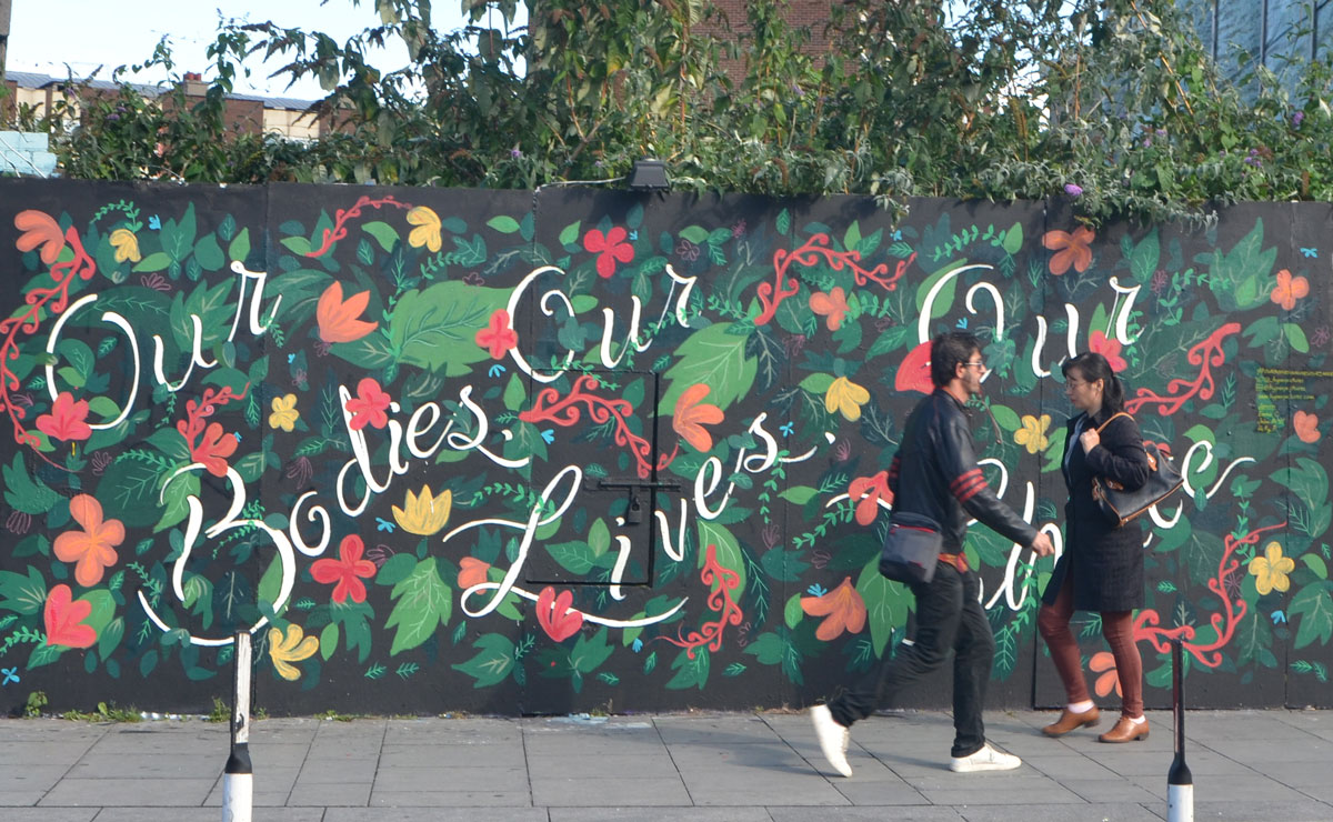 two people walking past a mural covered with pictures of flowers and leaves and the words, in cursive writing, Our bodies our lives our choice
