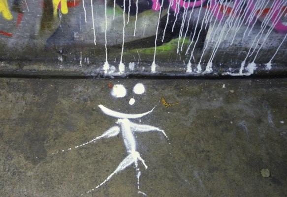 a happy face stick figure person made with paint that has dripped from a piece of street art, on a sidewalk (pavement)