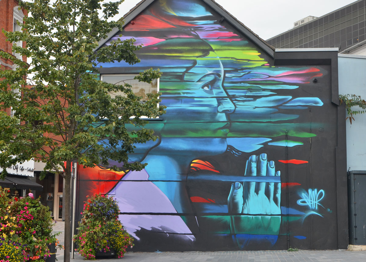 on the side of a two storey building, a large woman in profile, head and shoulders, in a multicoloured cloud, with her hands together in prayer, eyes open and looking slightly upwards