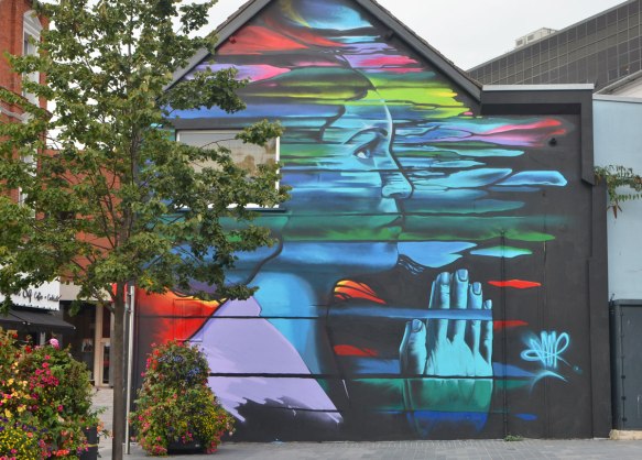 on the side of a two storey building, a large woman in profile, head and shoulders, in a multicoloured cloud, with her hands together in prayer, eyes open and looking slightly upwards